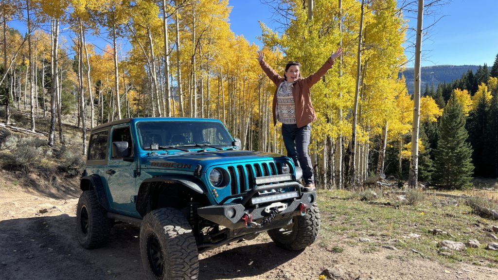 Melissa Cook stands on her blue jeep with fall colors behind her